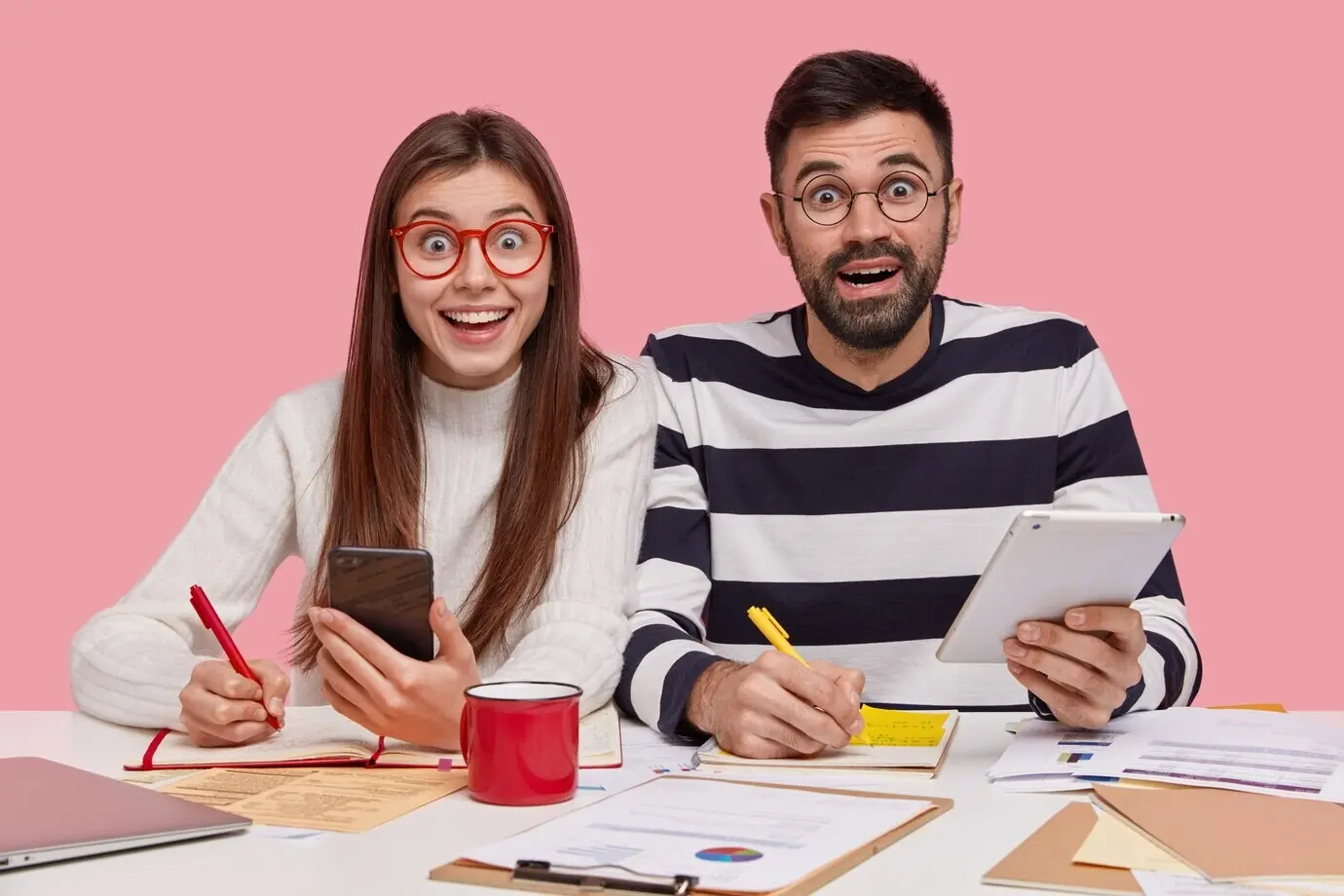 Team members sit at one desk, take notes in a notepad, prepare for a seminar, hold a modern smartphone and a touchpad, look happy, study documents with charts, and are engaged in the working process.